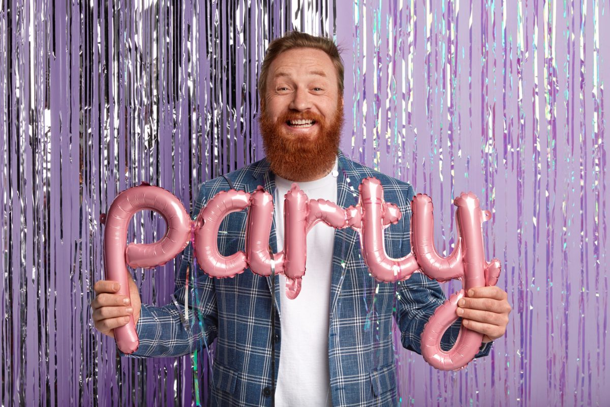 Horizontal shot of handsome man with thick red beard, poses with pink party balloons in form of letters, wears fashionable checkered jacket, has special event of festive occasion stands against tinsel